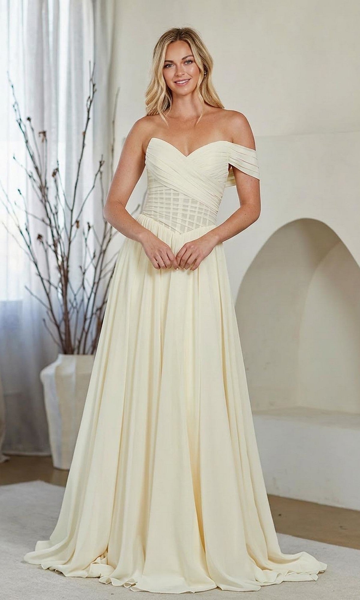 A woman stands indoors, smiling at the camera in the Amelia Couture 9097 long prom dress, featuring an ivory color, off-the-shoulder asymmetrical neckline, and fitted bodice.